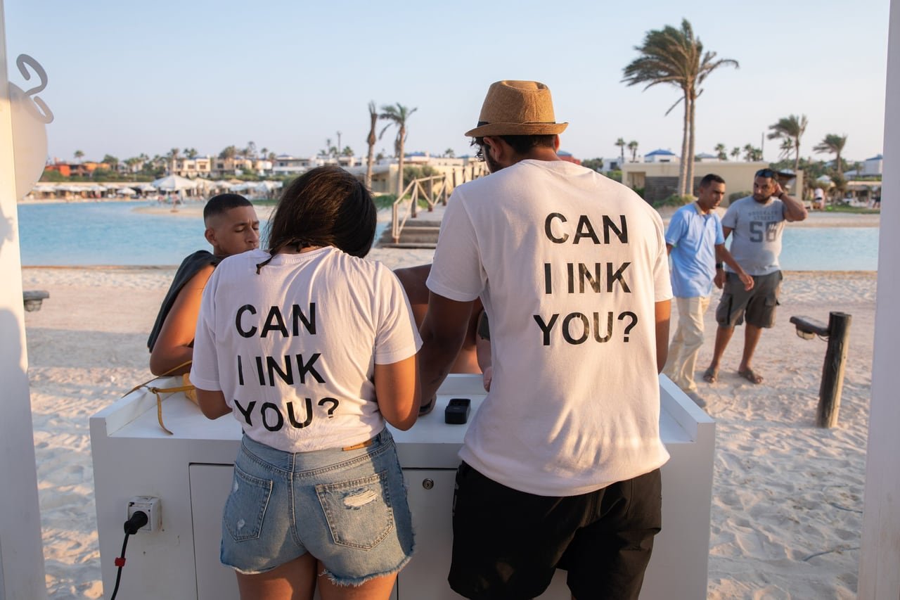 ضيفان بقمصان «Can I ink you?» عند ركن الوشم.